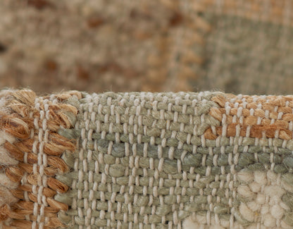 Close-up of a textured woven rug in sage green, ivory and tan with a tan fringe along the left edge.