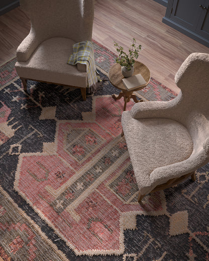 Two beige boucle armchairs with wooden legs, a round wood side table with plant and book, on a pink-navy geometric rug.