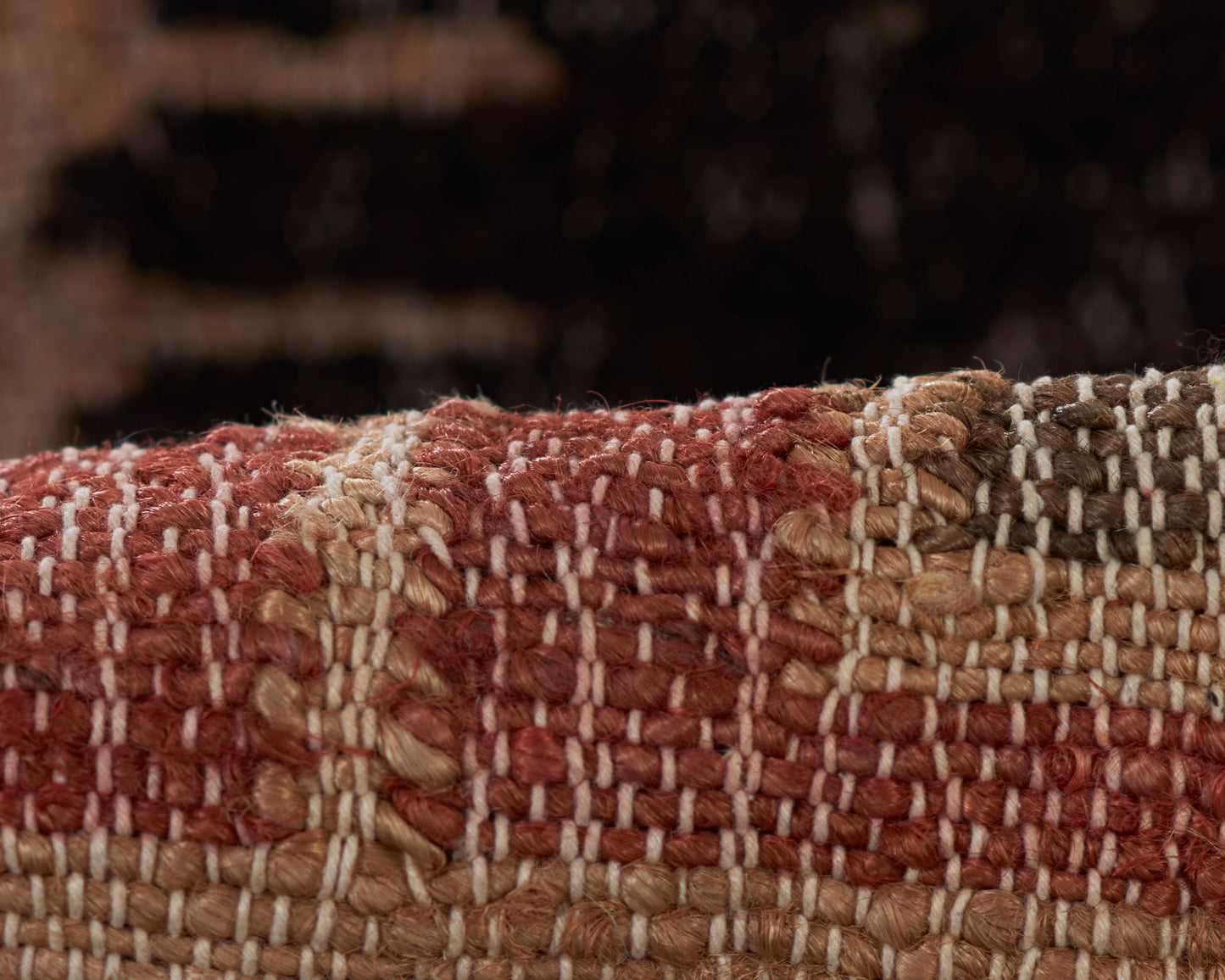 Macro shot of a woven rug with chunky red, tan and beige yarns in a checkered striped pattern.