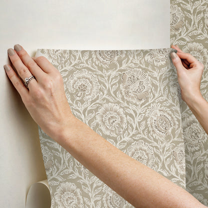Beige floral damask wallpaper sample with light gray background held by two hands.