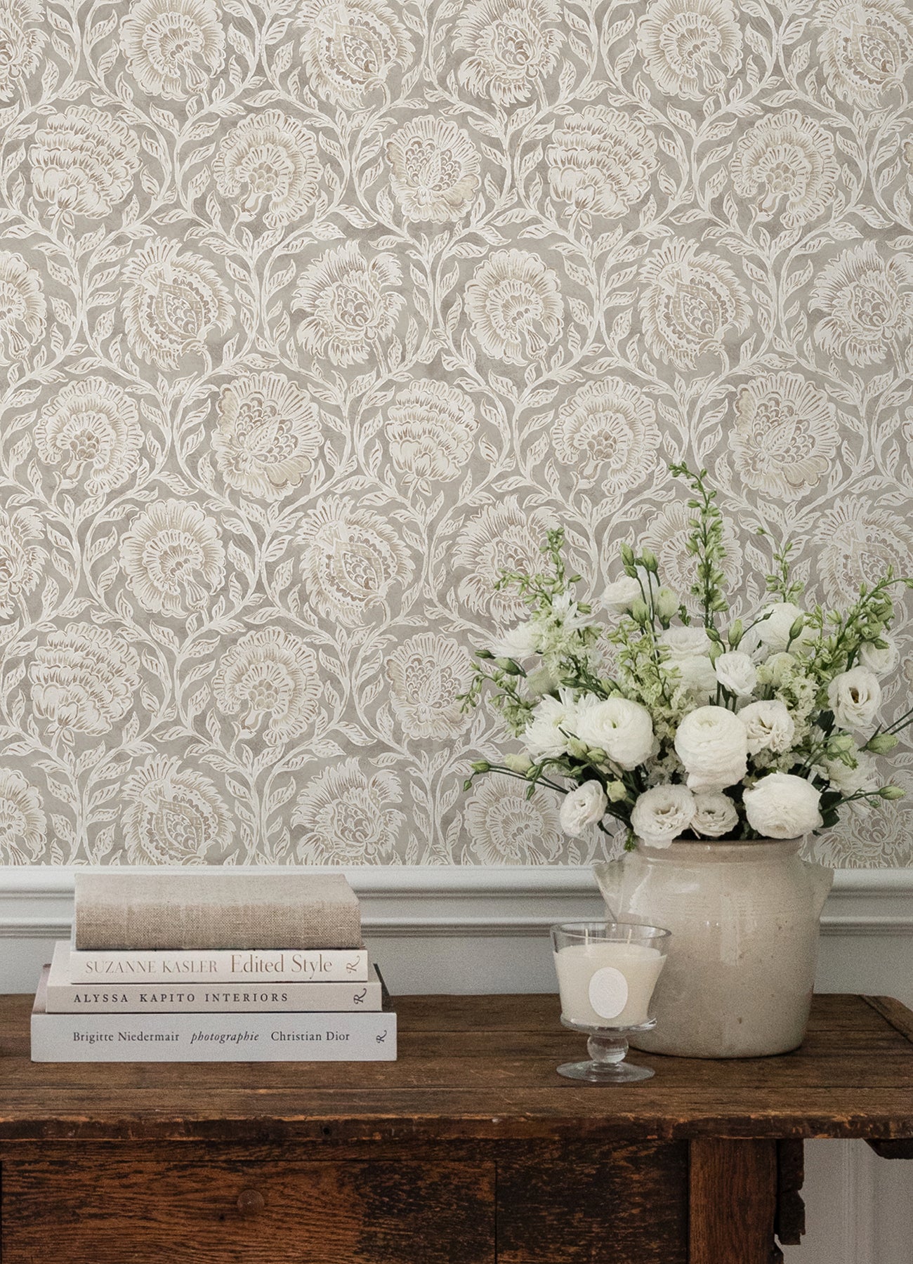 Beige damask floral wallpaper, rustic wooden table with stack of books, ceramic vase of white flowers, & a candle.