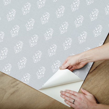 Light gray wallpaper with white floral motif; sample sheet peeled back to reveal white backing, hands shown.