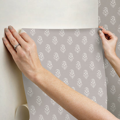 Hands hold a grey wallpaper sheet featuring a repeating white damask floral pattern.