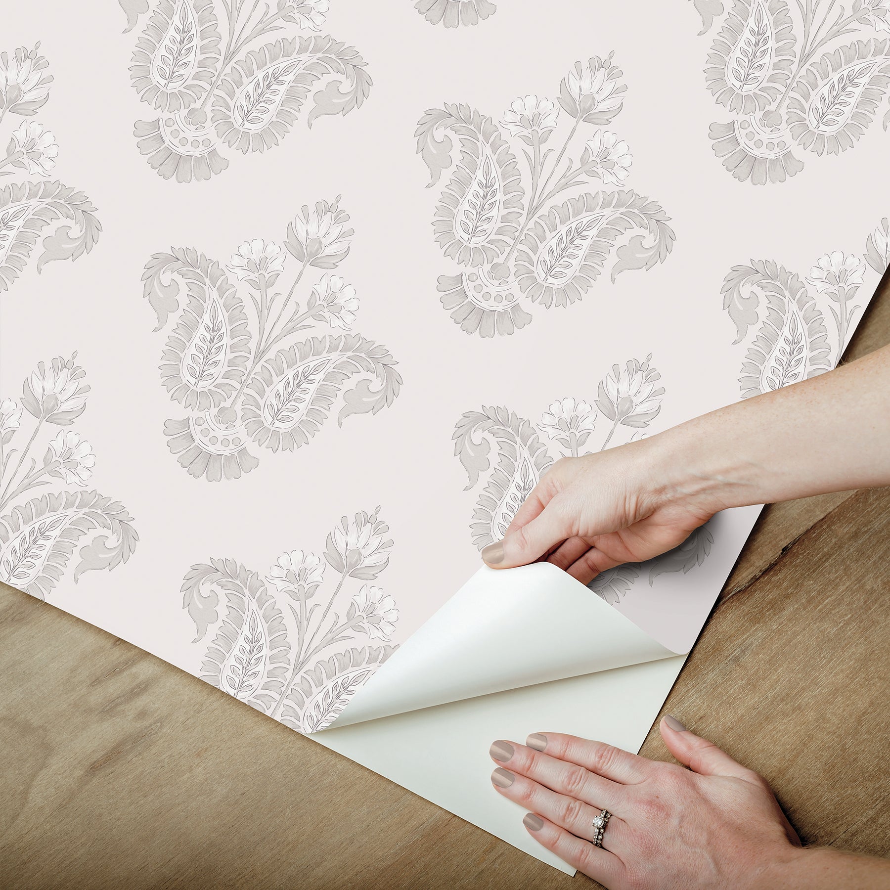 Hands peel back the corner of white wallpaper with a light gray damask pattern on a wooden floor.