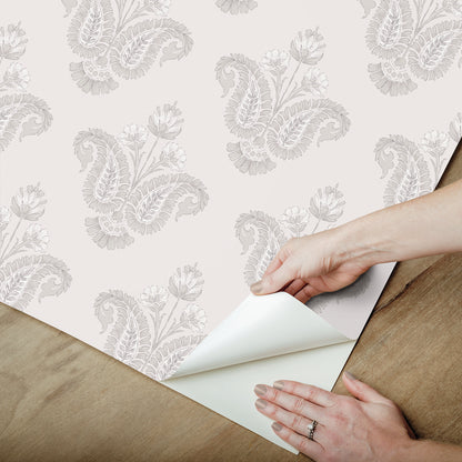 Hands peel back the corner of white wallpaper with a light gray damask pattern on a wooden floor.