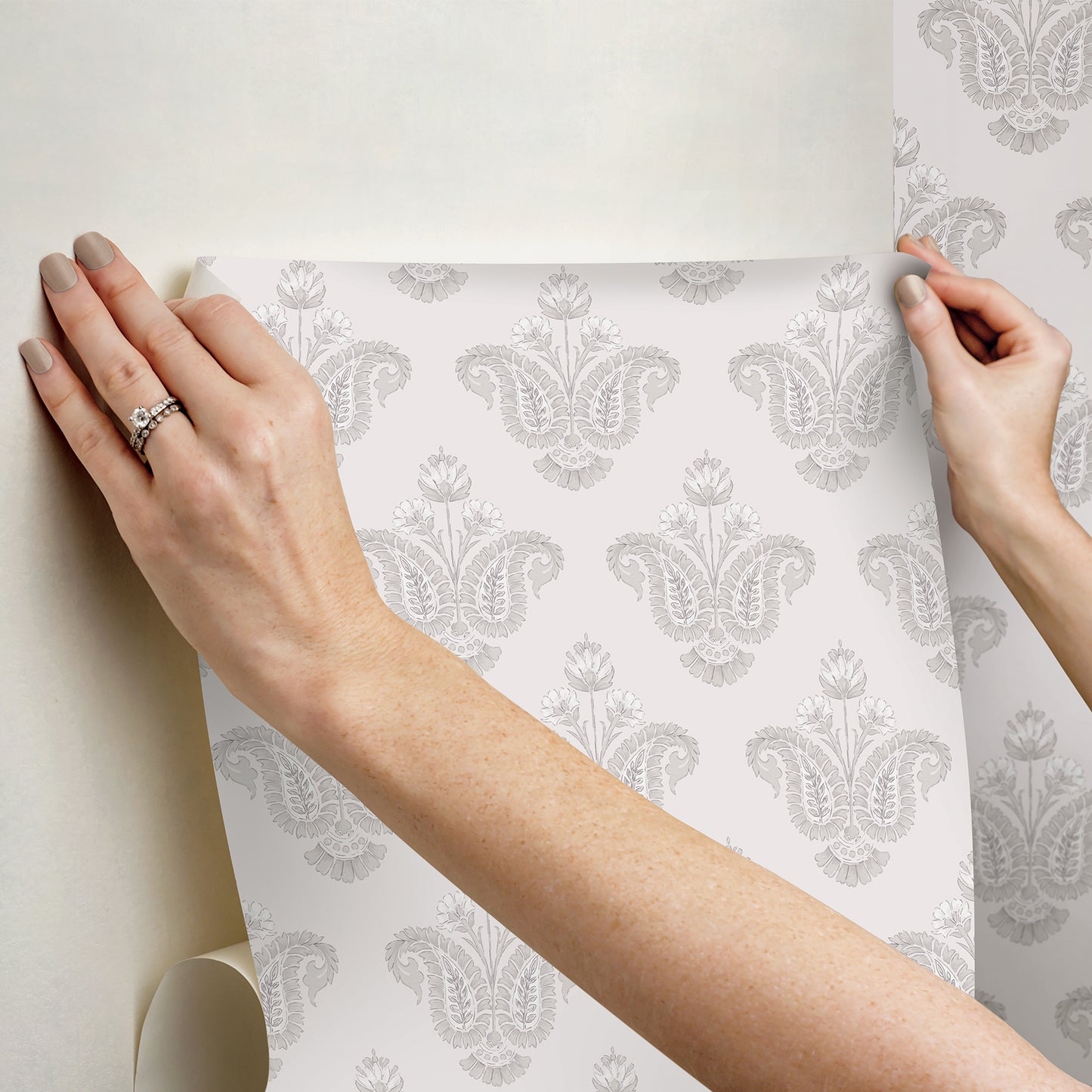 Hands smoothing a gray damask wallpaper sheet onto a light wall; decorative, classic pattern.