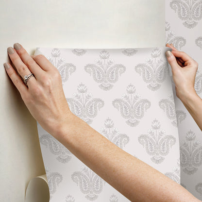 Hands smoothing a gray damask wallpaper sheet onto a light wall; decorative, classic pattern.