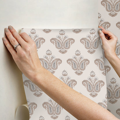 Hands apply a taupe damask wallpaper sheet to a cream background, showcasing an ornate vintage pattern for walls.