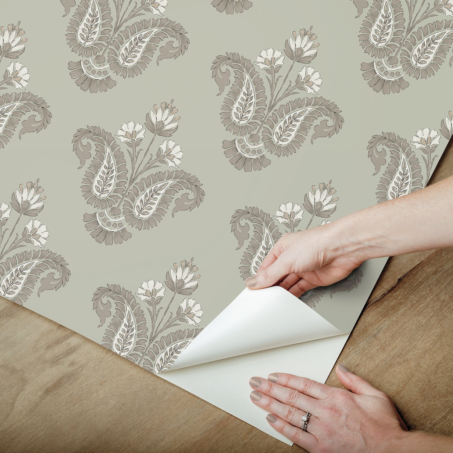 Hands peel back corner of taupe damask floral wallpaper on sage green background.