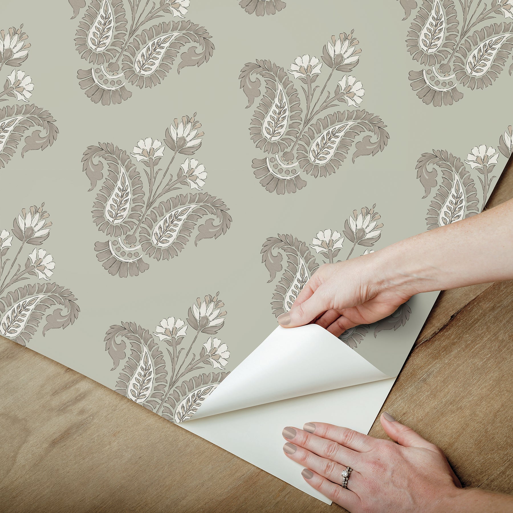 Hands peel back corner of taupe damask floral wallpaper on sage green background.