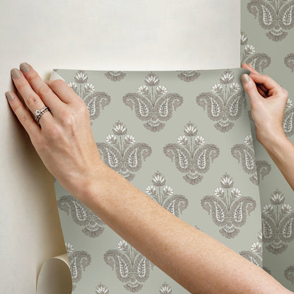 Hands hold a damask wallpaper sample with a pale blue-gray background and ornate gray pattern.
