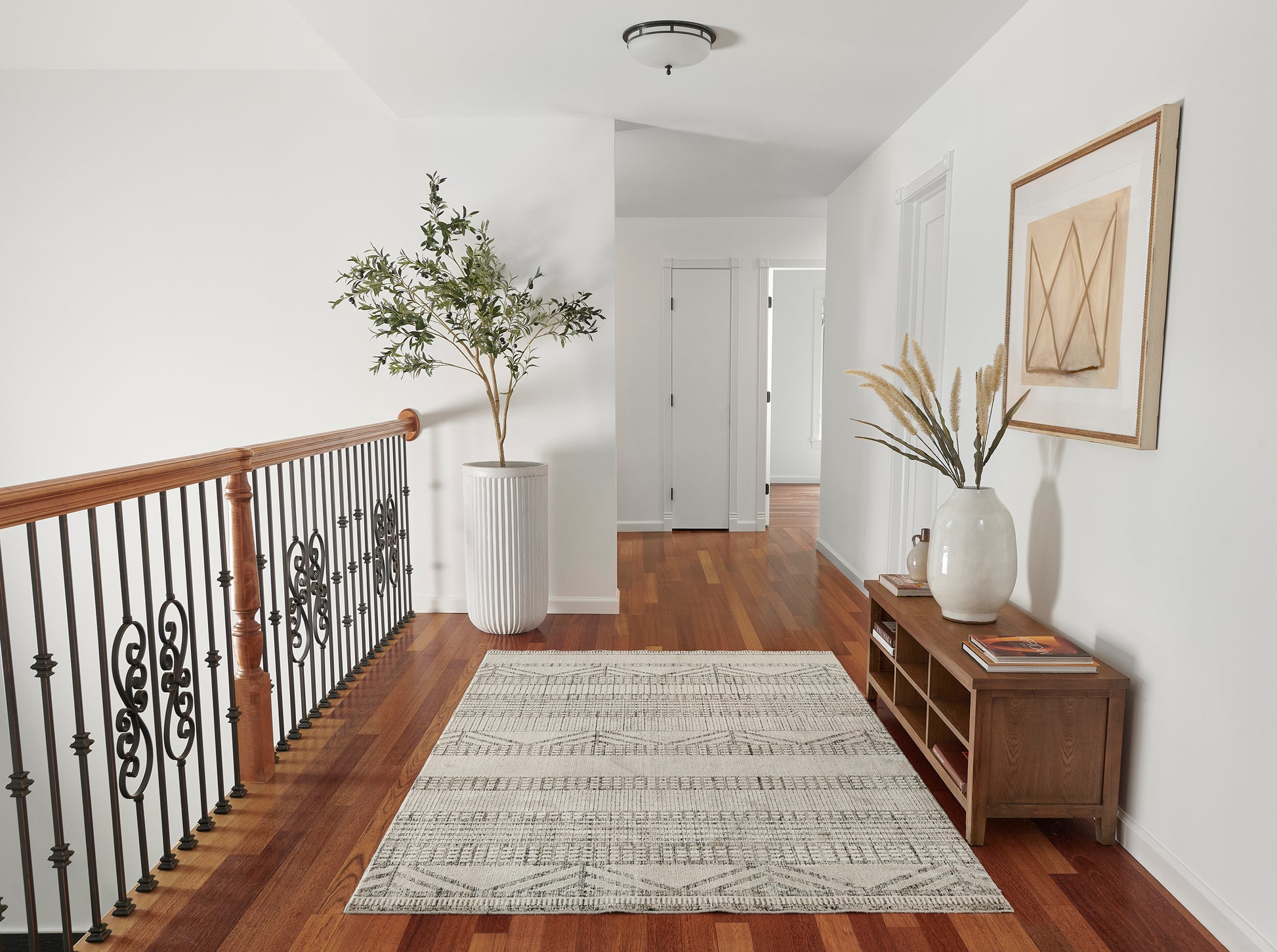Bright hallway with warm wood floor, black iron railing, tall white planter, gray rug, wooden console with vase.
