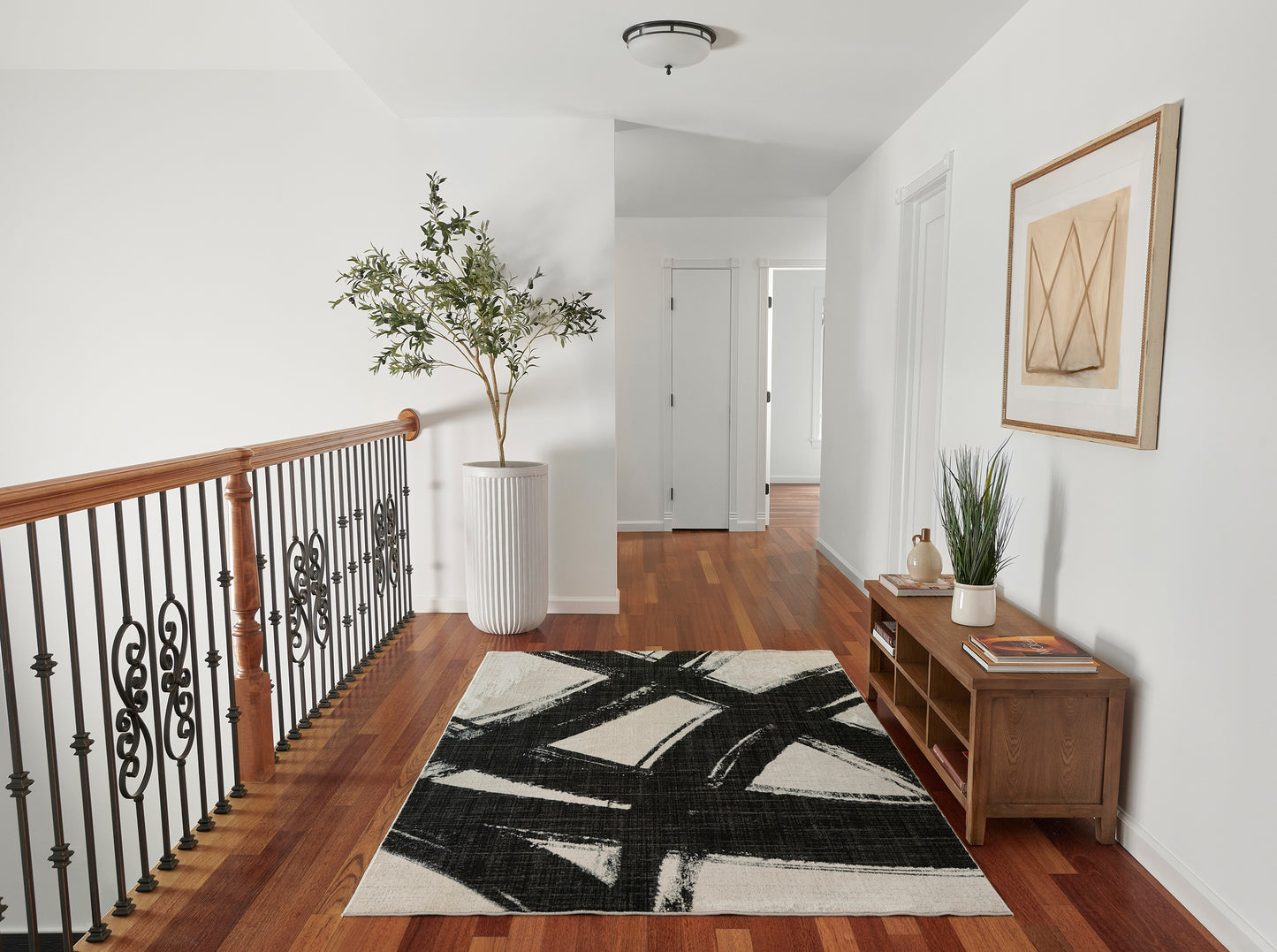 Bright hallway with wood floor, black-and-cream abstract rug, wooden console with plant and books, framed art, iron railing.