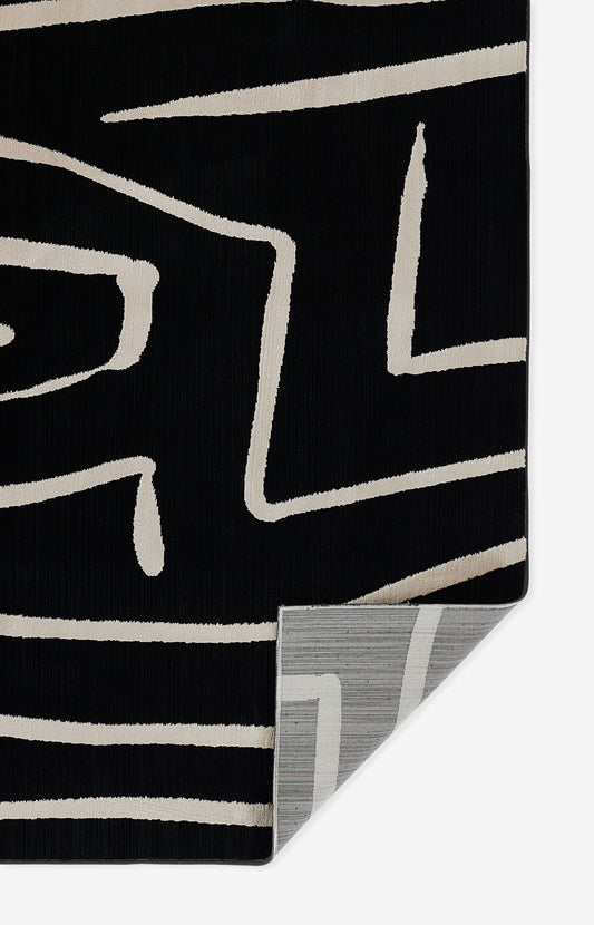 Black rug with beige abstract geometric lines; thick pile, folded corner reveals gray striped backing.