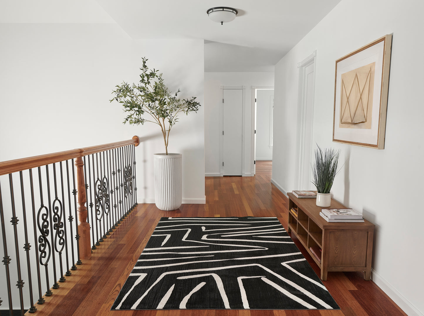 Hallway with black geometric rug on wood floor, wrought-iron railing, tall potted plant, wooden console with mags & plant, framed art.