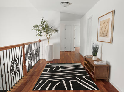 Hallway with black rug featuring white geometric lines, warm wood floors, iron railing, tall potted plant, and wooden console.
