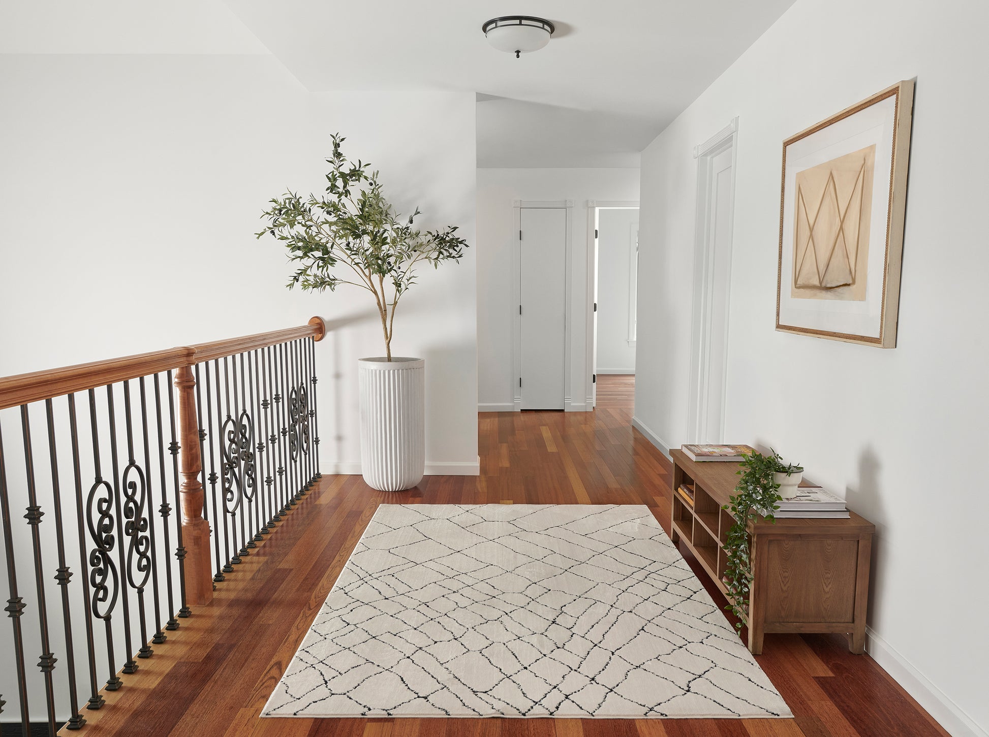 Tall leafy plant in a white ribbed planter beside a wooden railing, beige geometric rug, wall art, and a console with books.