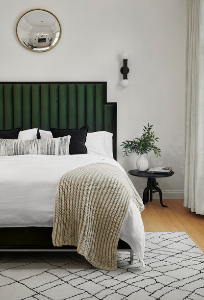 Emerald green velvet headboard bed with white linens, beige knit throw; gold mirror, black wall sconce, plant on side table.