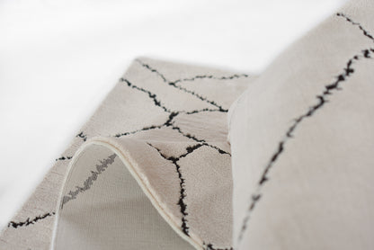 Beige rug with black geometric lattice pattern, soft textured pile, rolled edge revealing underside.