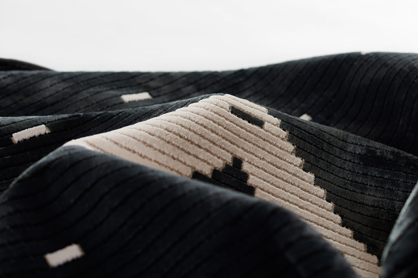 Close-up of a black corduroy rug with a beige ribbed panel and small white patches; textured, cozy rug.