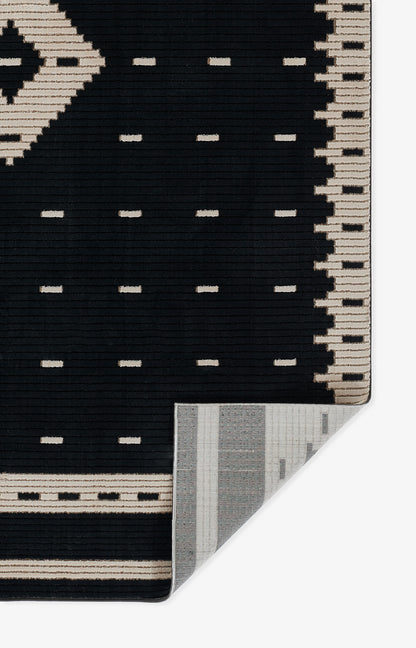 Black rug 9' x 13' with beige geometric border and small beige dash accents; folded corner reveals grey striped underside.