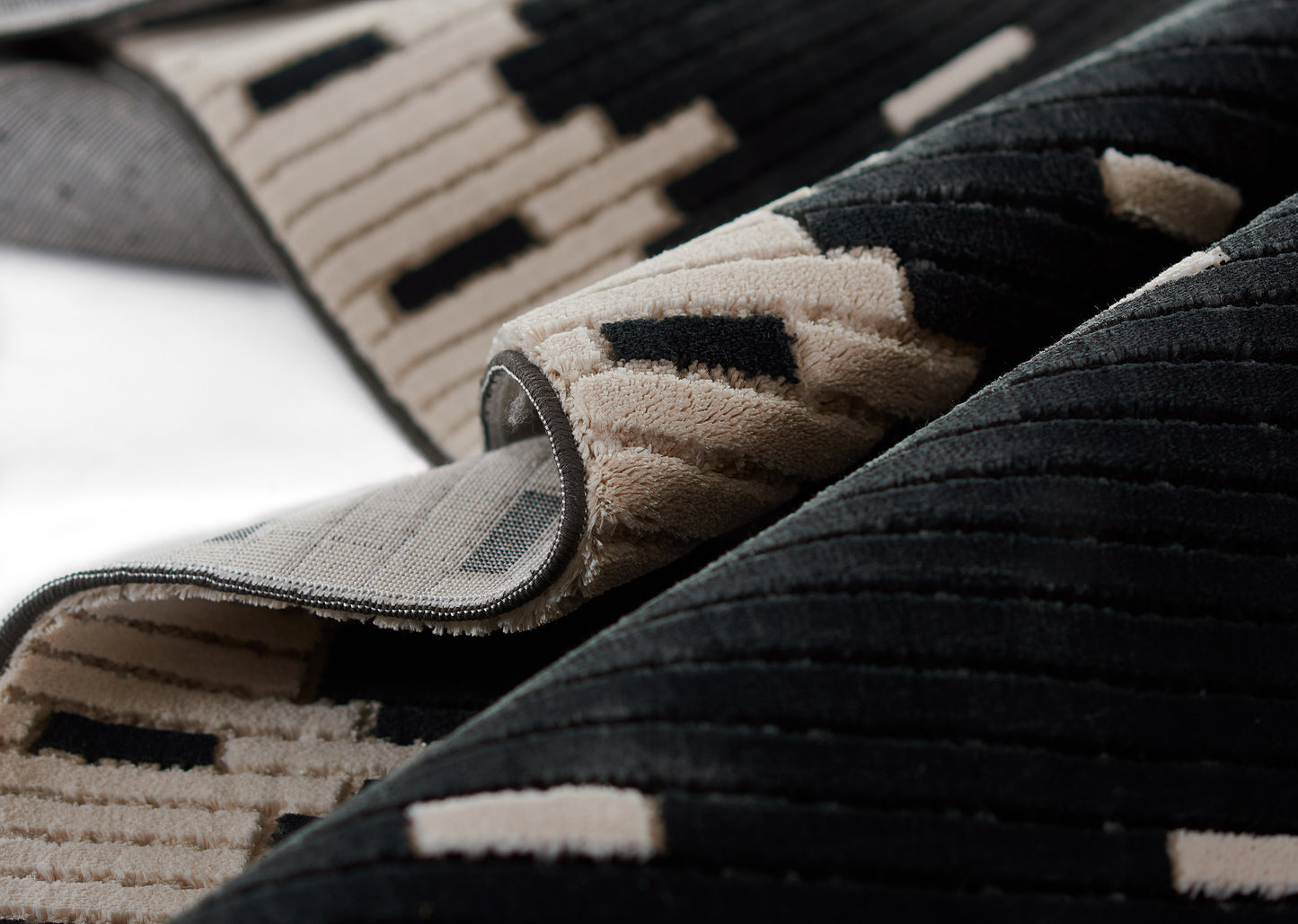 Beige and black geometric plush rug, folded to reveal grid-patterned underside and thick stitched edge.