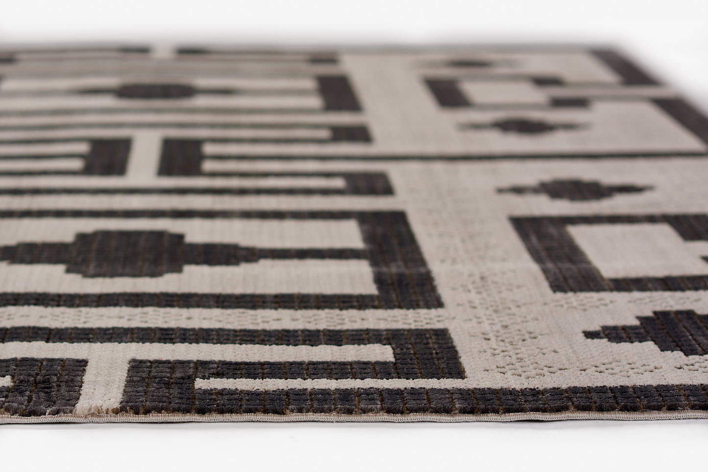 Beige woven rug with dark brown abstract geometric pattern, low-pile texture and light fringe along edge.