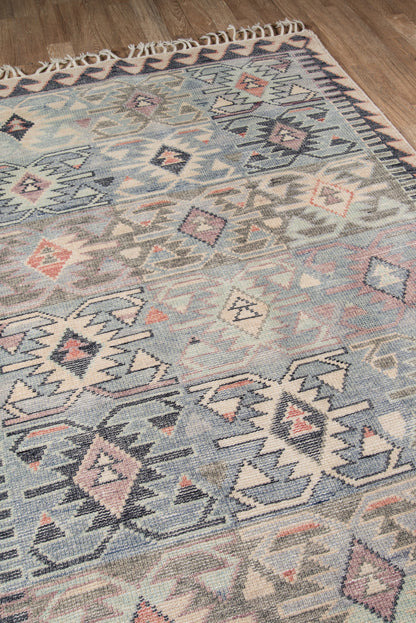 Blue geometric tribal rug with ivory, gray and pink motifs, fringed edges on a wood floor.
