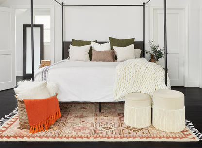 Geometric rug on floor in a bedroom setup; black four-poster bed with white linens, tan/cream pillows, chunky knit throw, orange throw on a wicker basket, and cream ottomans.