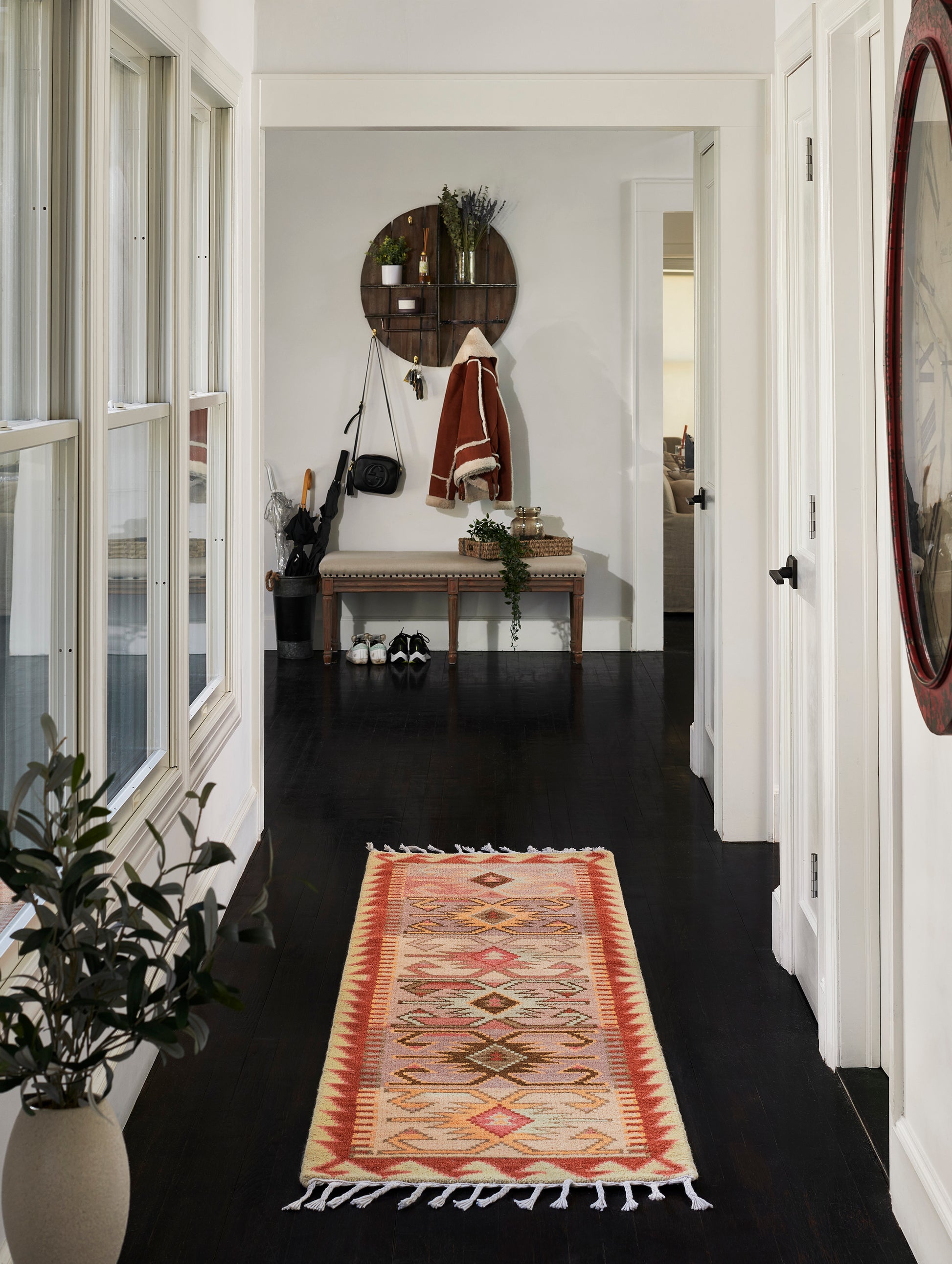Geometric warm-red cream runner rug in a dark hallway with a bench, coat rack, round shelf, plants, and a red-framed mirror.
