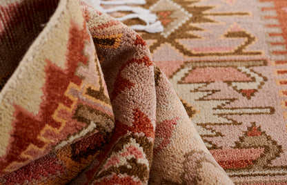 Close-up of pink, beige and brown tribal rug with geometric motifs, plush tufted pile, folded corner and white fringe.