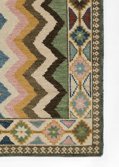 Nomad-style wool area rug with bold chevron zigzags in green, cream, brown, pink and blue; ornate bordered motif.