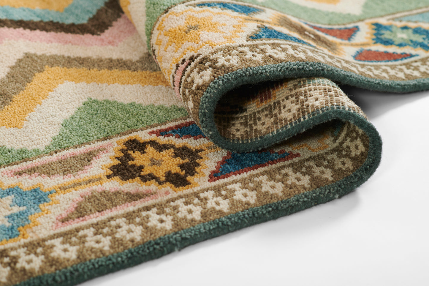 Colorful tribal rug with geometric motifs, rolled corner, turquoise edging, pink, green, beige, brown hues.