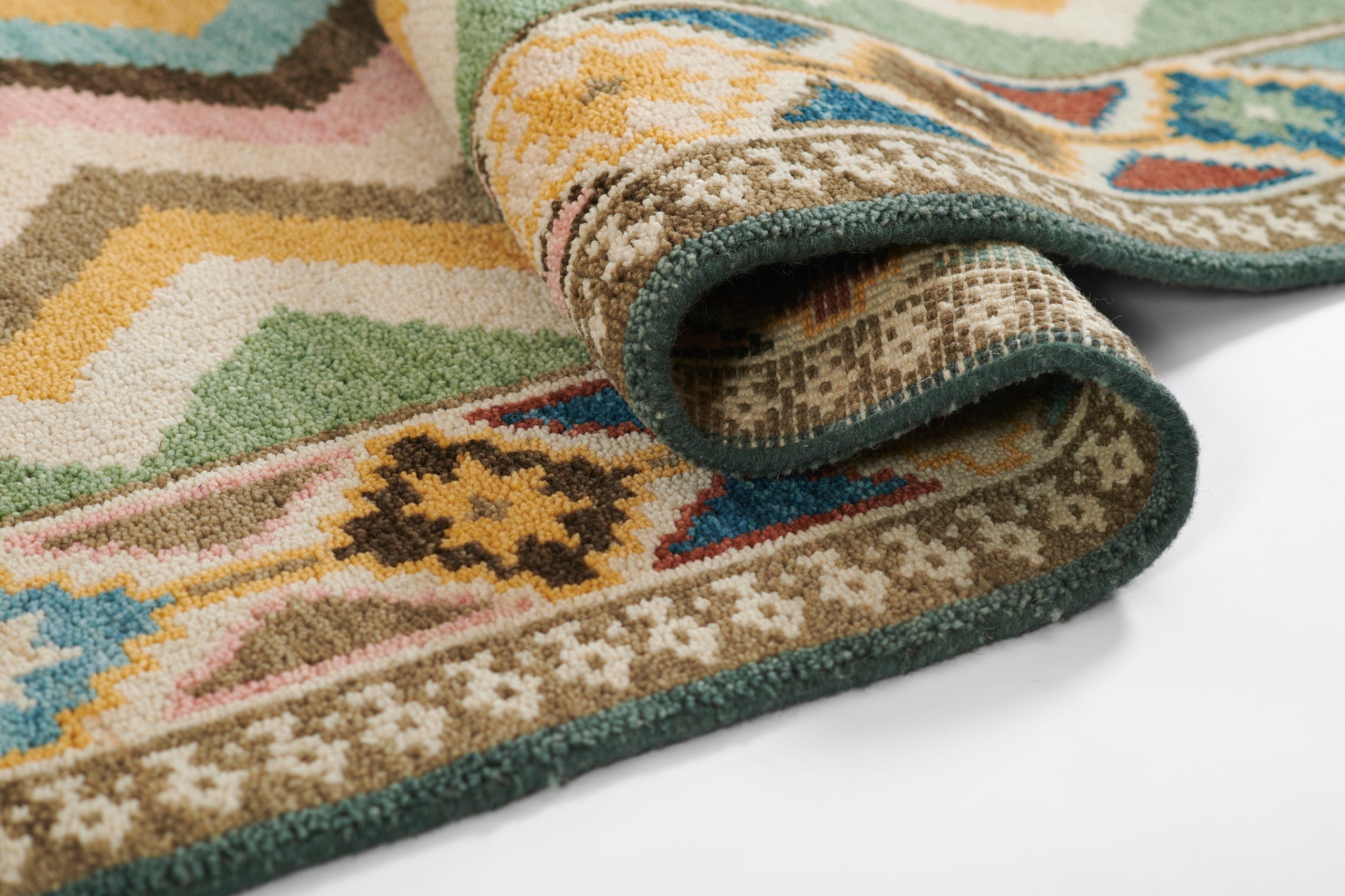 Colorful tribal rug with geometric motifs, rolled corner, turquoise edging, pink, green, beige, brown hues.