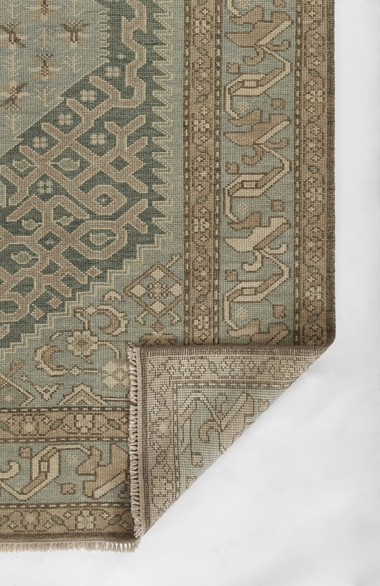 Nomad-style rug in muted teal, taupe and beige with geometric pattern and decorative border; folded corner shows backing.