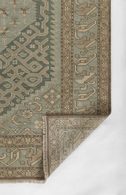 Nomad-style rug in muted teal, taupe and beige with geometric pattern and decorative border; folded corner shows backing.
