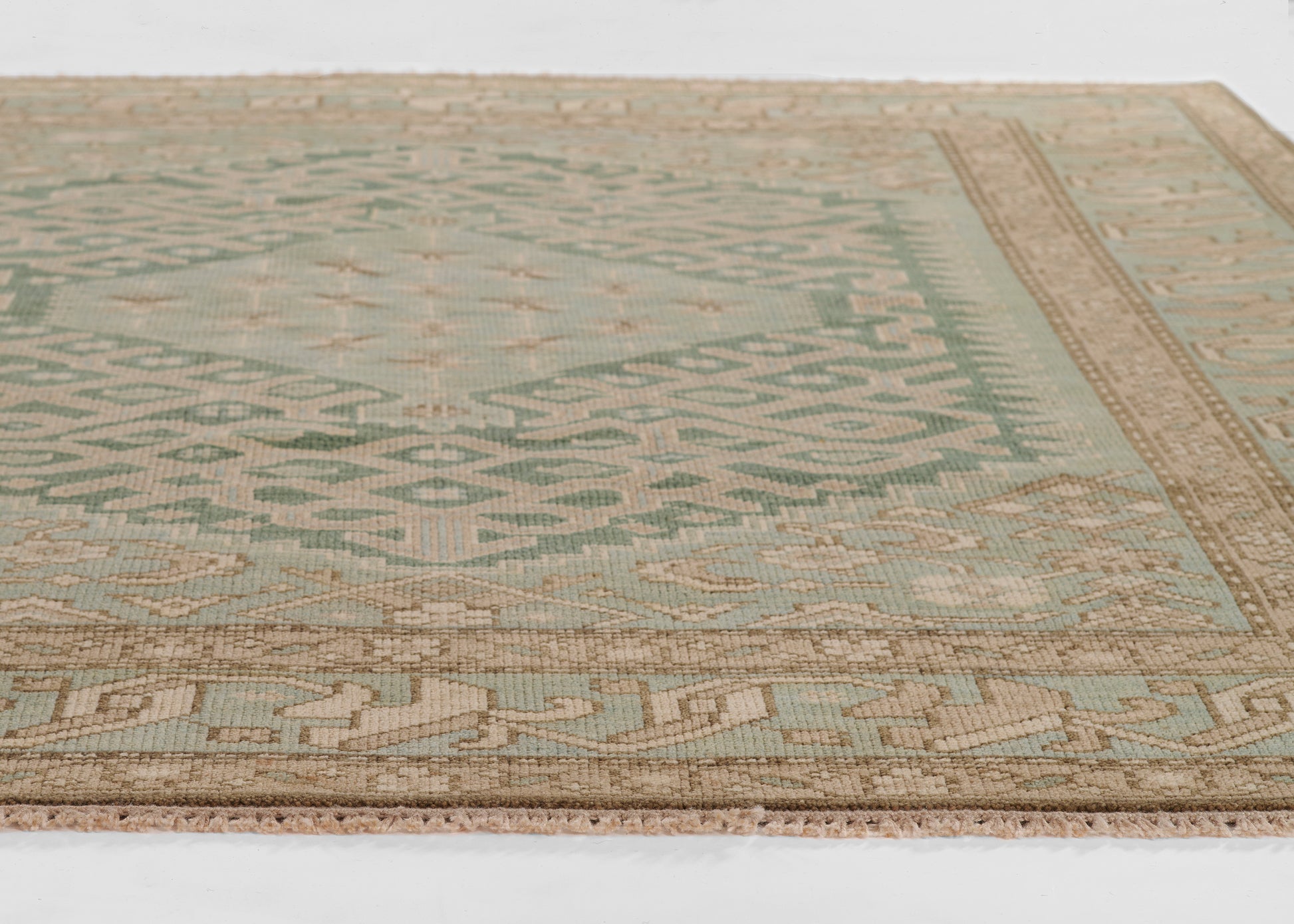 Nomad-inspired flat-weave rug in sage green and beige with geometric motifs and tan bordered edges.