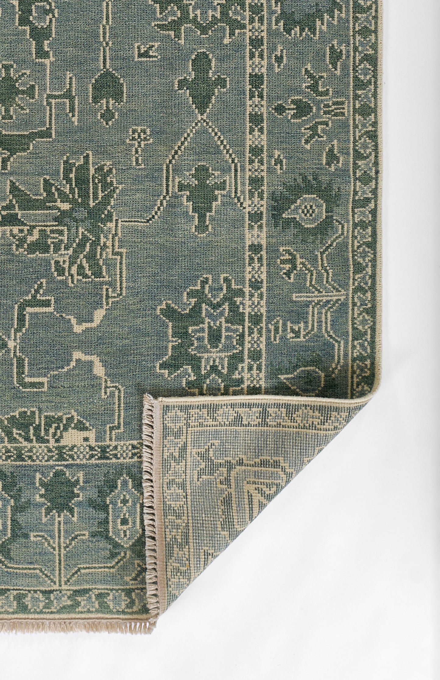 Blue-green nomad-style woven rug with cream geometric motifs, bordered edges and beige fringe at a folded corner.