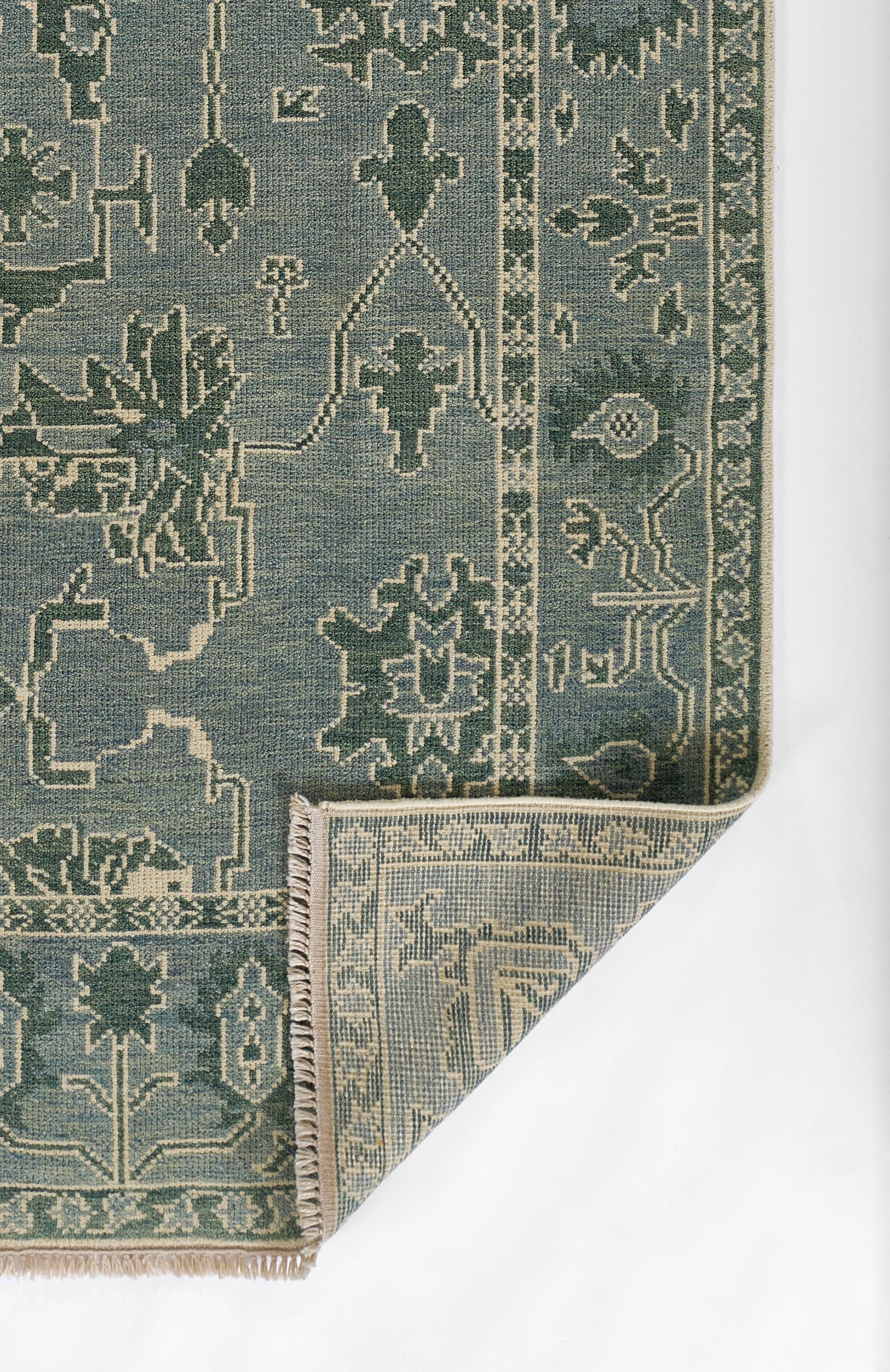 Blue-green nomad-style woven rug with cream geometric motifs, bordered edges and beige fringe at a folded corner.