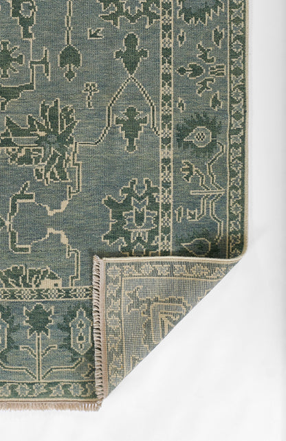 Blue-green nomad-style woven rug with cream geometric motifs, bordered edges and beige fringe at a folded corner.