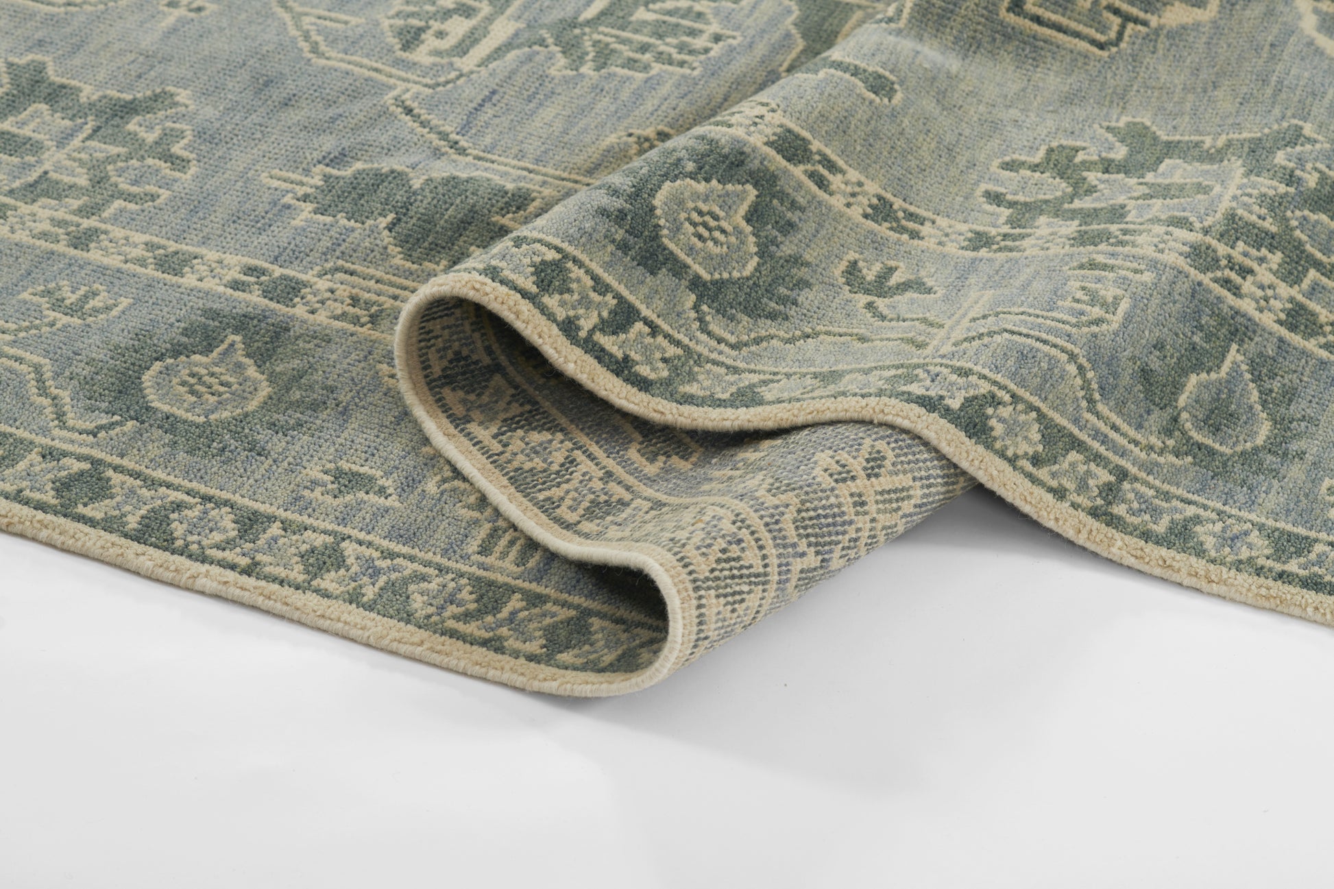 Blue-green oriental rug with cream fringe, intricate floral border; rolled corner shows soft woven texture.