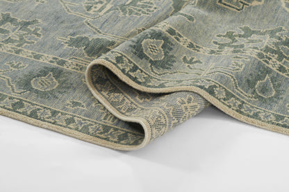 Blue-green oriental rug with cream fringe, intricate floral border; rolled corner shows soft woven texture.
