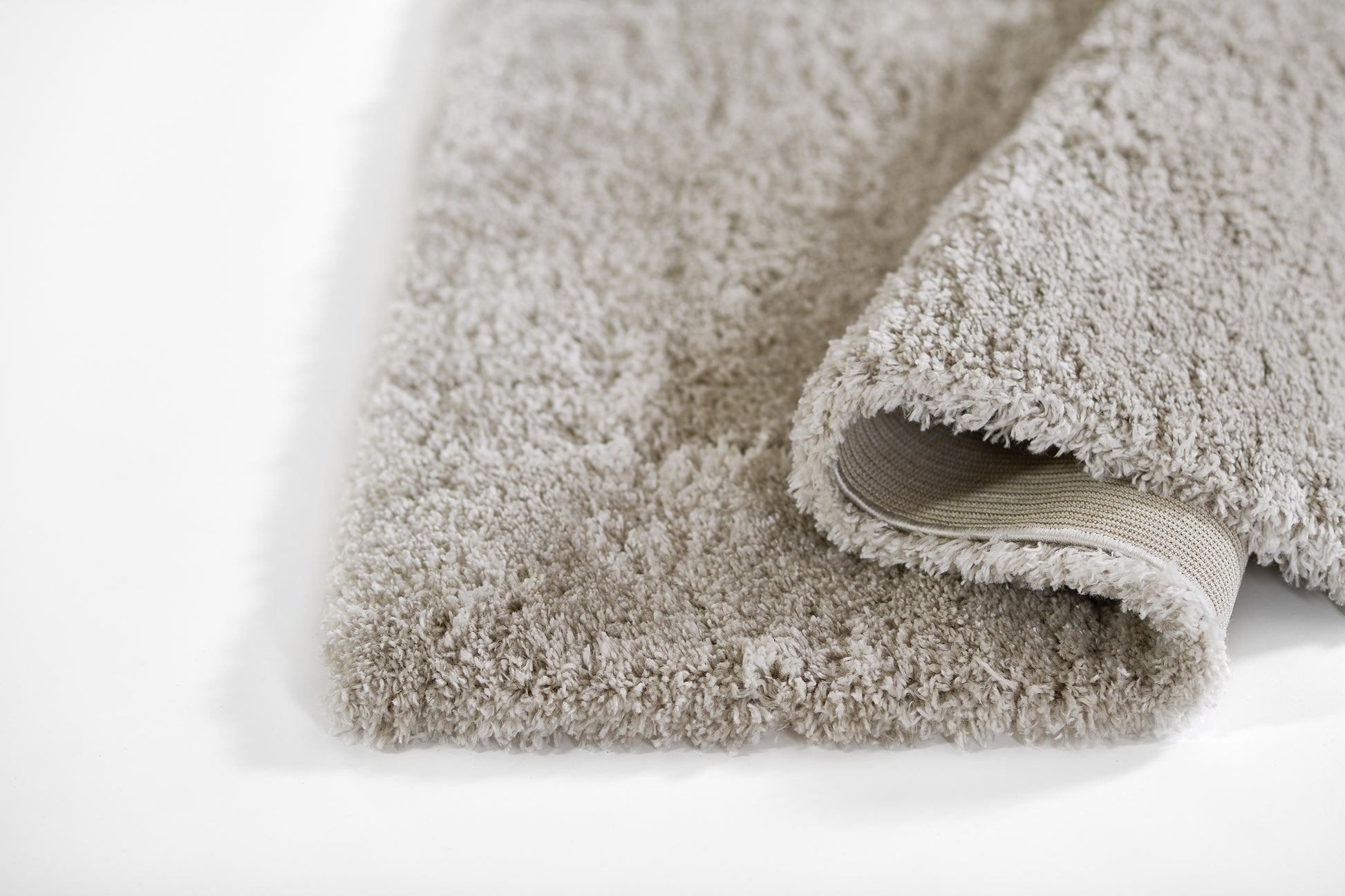 Beige shag rug with plush pile, thick rectangular area rug, rolled edge reveals non-slip backing; cozy floor mat.