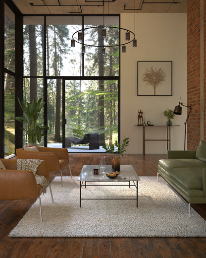 Modern living room with large forest-view windows, tan leather chairs, green sofa, glass coffee table on white rug.