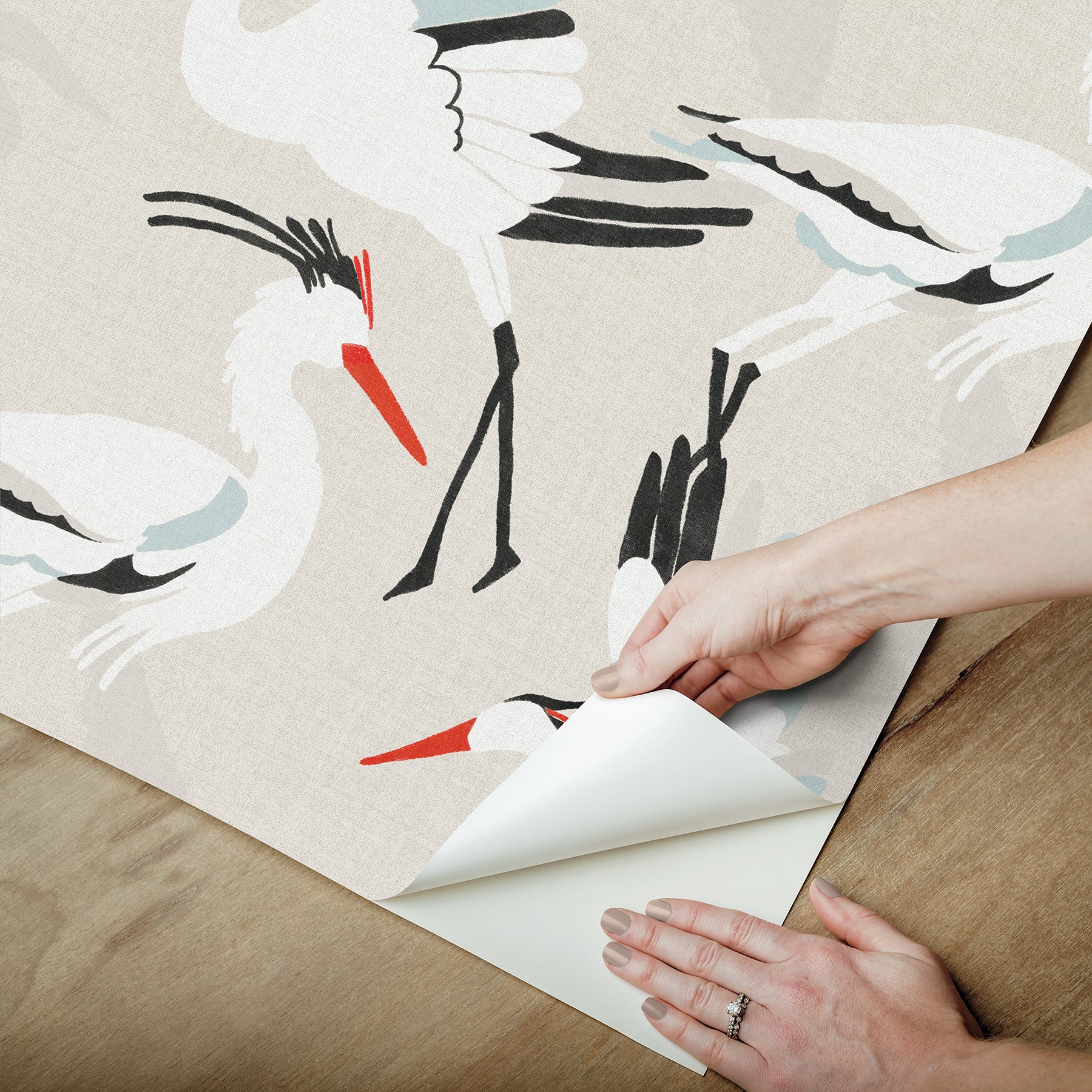 Beige wallpaper featuring white cranes with red beaks; a hand peels back the backing from the corner.