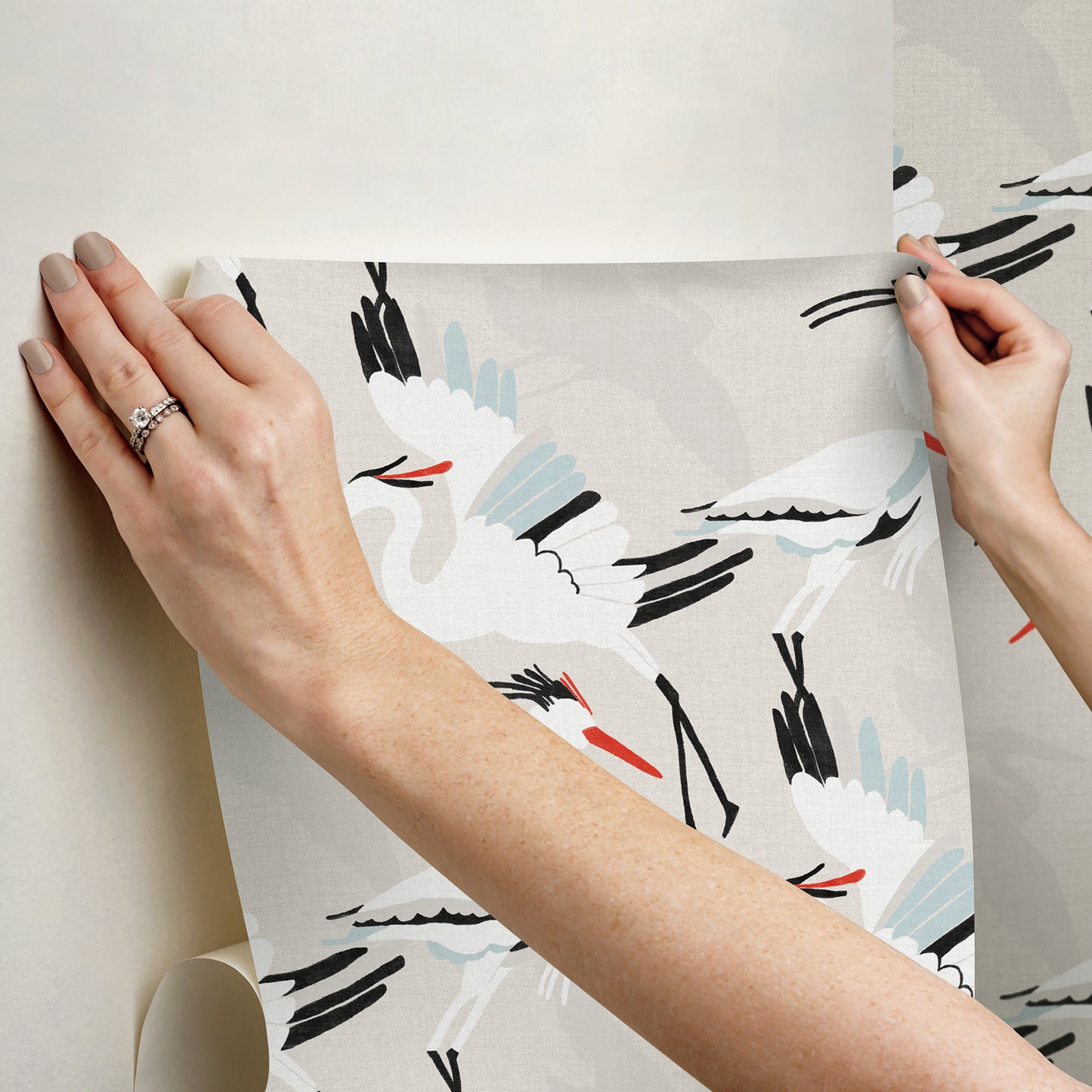 Two hands hold a sheet of light grey wallpaper with white cranes, black accents & pale blue highlights.