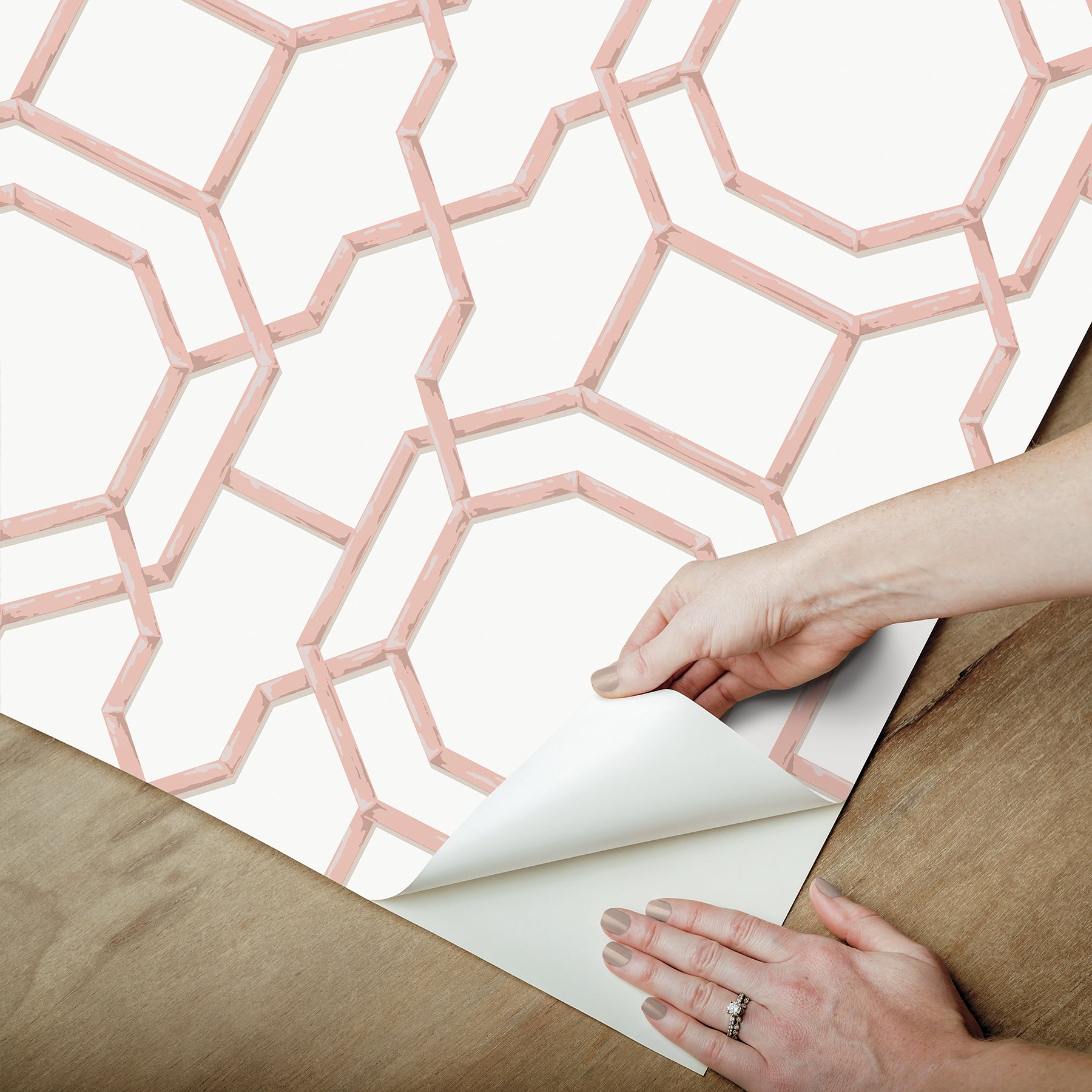 Pink geometric lattice wallpaper sheet being peeled back to reveal white backing on a wooden table.