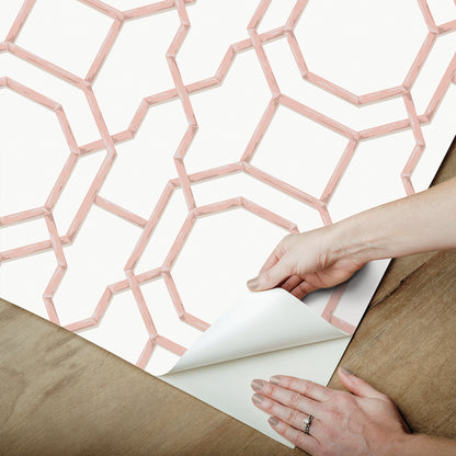 Pink geometric lattice wallpaper sheet being peeled back to reveal white backing on a wooden table.