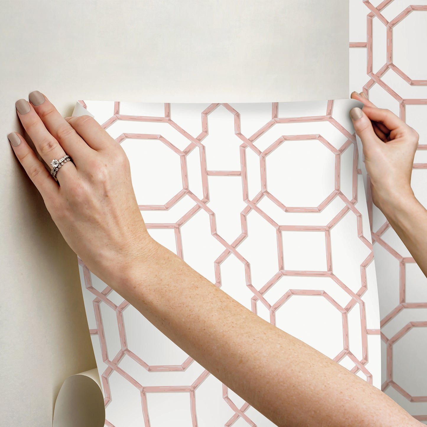 Hands hold pink geometric lattice wallpaper against white background; modern decorative wallcovering.