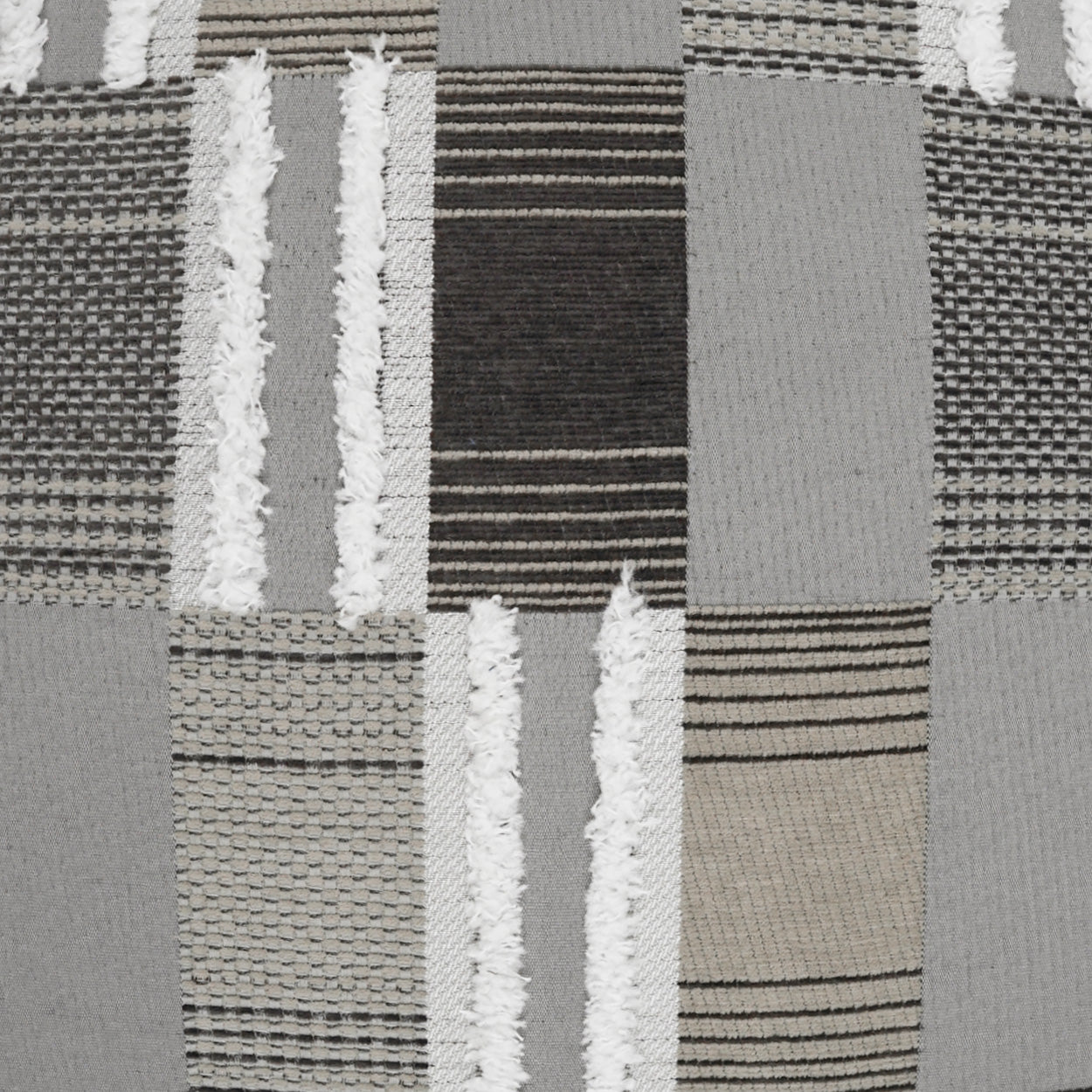 Outdoor pillow with patchwork grey, black, and beige woven fabric featuring striped and textured blocks in a quilt-like pattern.